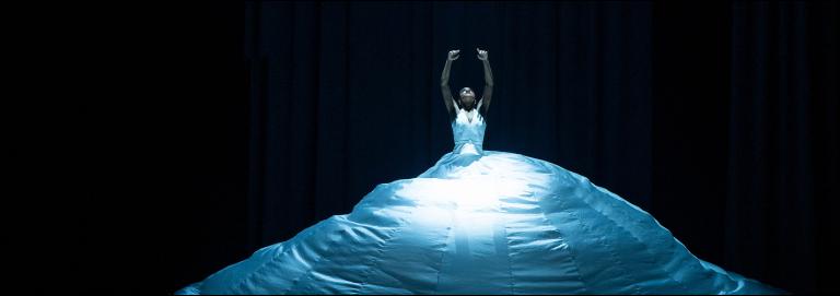 Dancer wearing a massive skirted gown looks up with her arms raised up