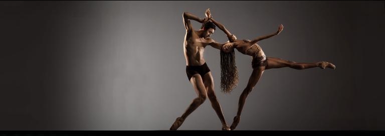 Two dancers wearing brown and black appear in front of a gray backdrop. One dancer extends a leg straight to the right while bending backward toward the second dancer, who stands at a slight angle with one leg bent at the knee, holding onto the arm of the other dancer.