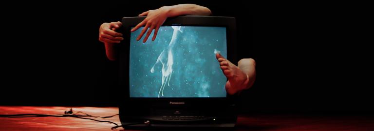 A black & white television is being embraced from behind and has an image of a hand reaching on the screen