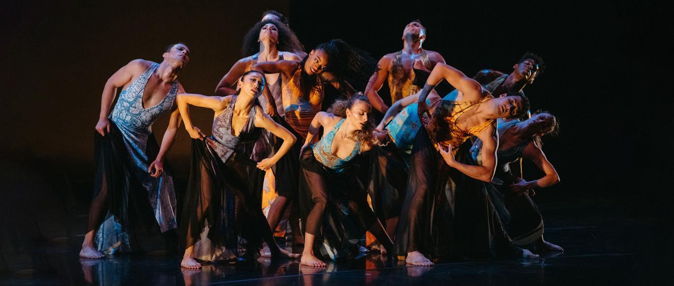 A group of nine dancers all wearing an assortment of shades of blue and orange, are huddled together center stage, each posing with their torso bent towards the audience, and their hands on their hips.