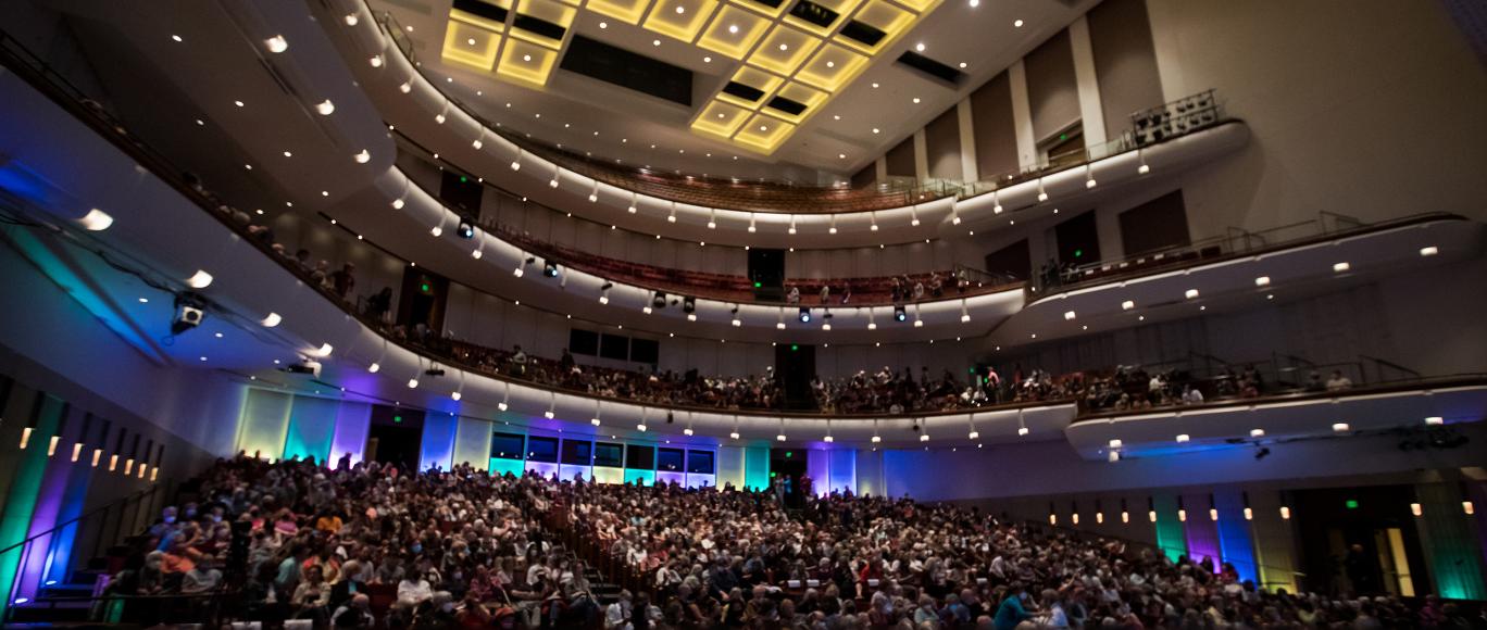 From the view of being on stage, the seats of a large, four level auditorium are lit with blue, purple, yellow, and green colored lights.