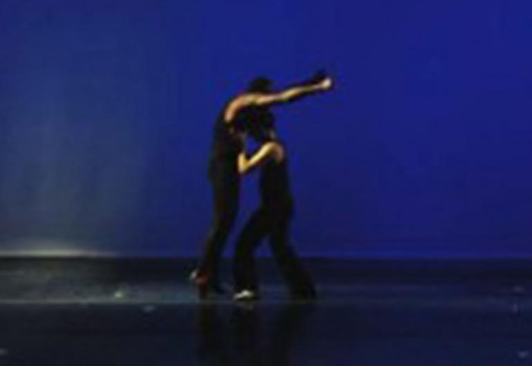 Video still of 2 dancers, one with head on the chest of the other whose arms are extended over them.