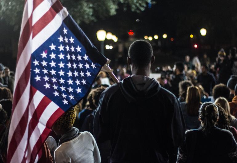 A crowd of people are outside at night with their backs facing the camera. A person close to the camera is holding a flag pole with an American Flag.
