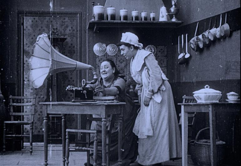 Black and white still of a woman at a desk with a gramophone and another woman in maid's uniform standing behind.
