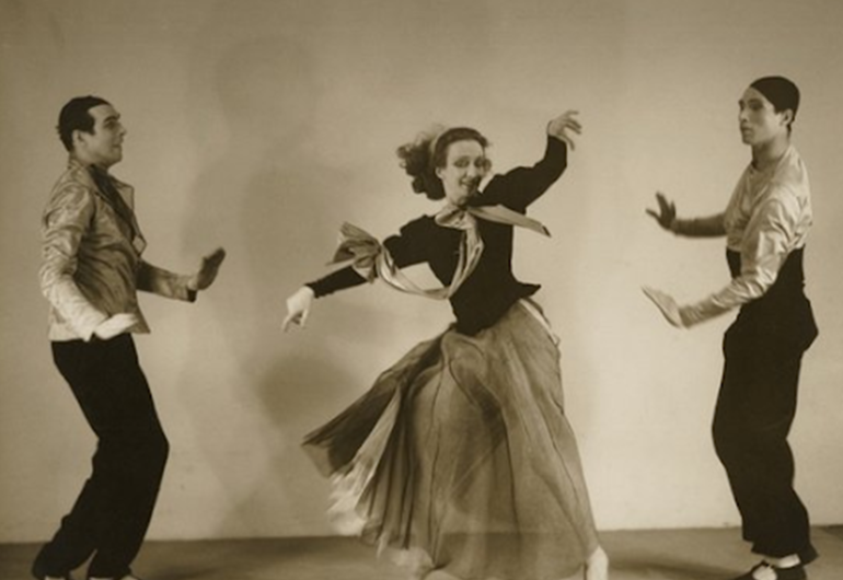 A sepia toned image shows three dancers. The dancer in the middle is wearing a long dress where the bottom is lighter than the top. Their arms are out to the side with the dancer’s left arm slightly raised above their right. There is one dancer on each side of the dancer in the dress. They both are wearing dark pants and a lighter top. They are faced toward the dancer in the middle.