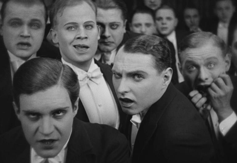 A group of men in tuxedos with varying expressions on their faces from shock, amusement, and disgust.