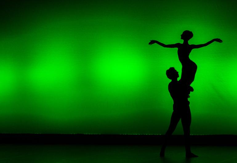 A silhouette of a dancer carrying another dancer by their legs appears in front of a green background. The lifted dancer's arms are a straight line. 