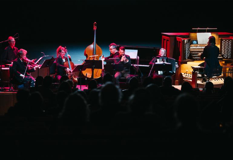 An ensemble of 7 musicians including a drum player, violinist, bassist, cellist, horn player, pianist, accordionist, and pipe organist dressed in black perform onstage with a dark background and pink lighting. 