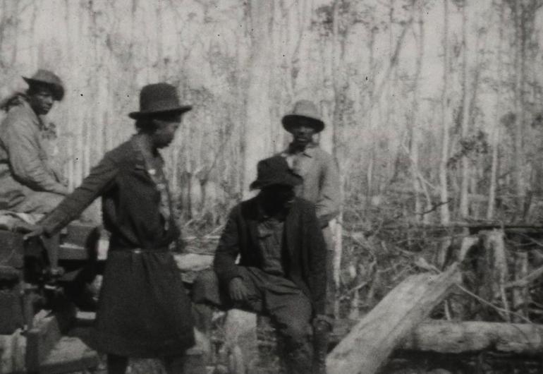 A black and white image shows 4 figures wearing hats, coats, and pants grouped within the left of the frame. They stand outside with trees surrounding them.