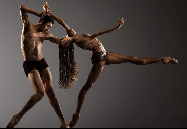 Photo © RJ Muna. Two dancers wearing brown and black appear in front of a gray backdrop. One dancer extends a leg straight to the right while bending backward toward the second dancer, who stands at a slight angle with one leg bent at the knee, holding onto the arm of the other dancer.