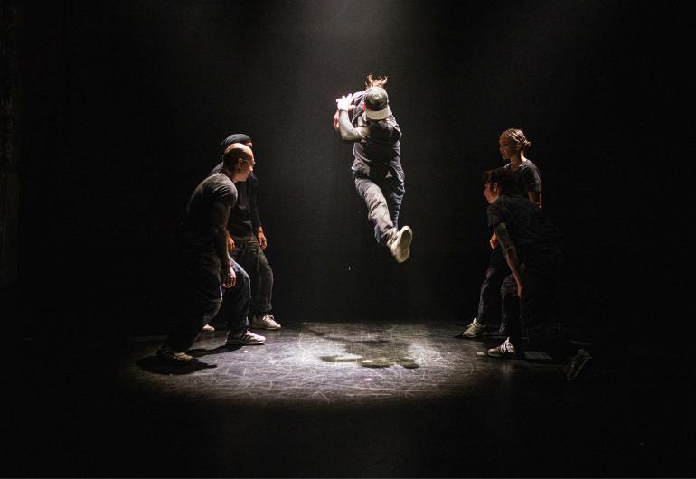 Five dancers appear in a spotlight otherwise surrounded by darkness. Four of the dancers form a circle around the fifth dancer who is up in the air with their leg outstretched.