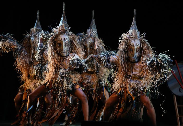 A group of dancers wearing creature-like costumes with pointed headwear appear onstage, facing the camera with their legs bent out toward their sides.