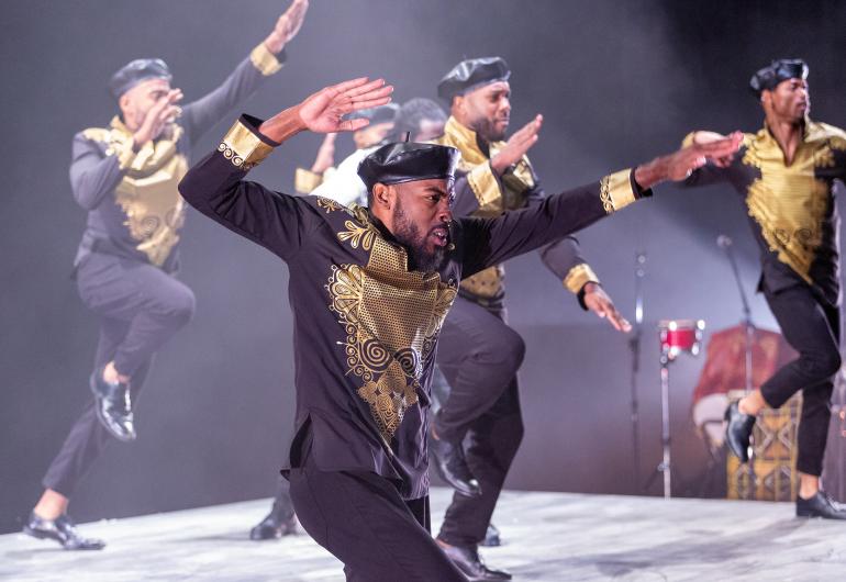 A group of dancers wearing black and gold clothing and caps appears onstage, jumping toward the right of the frame with one hand raised and bent behind them and the other pointed out.