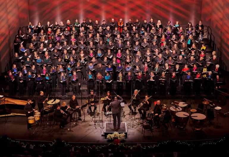 A choir wearing black with various colored scarves performs onstage with patterned red lighting projected behind them. A conductor stands downstage with their back toward the camera and musicians surrounding them.