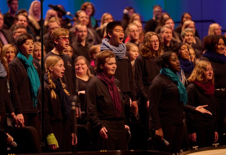 A group of chorus singers dressed in black with a mix of colored scarves perform with their hands resting to their sides, lined up in verticle rows of increasing height.