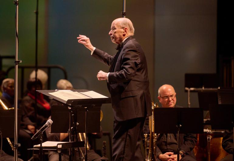 A conductor dressed in black faces to the left with one arm lifted and the other bent at their side. A music stand and musicians appear below them.