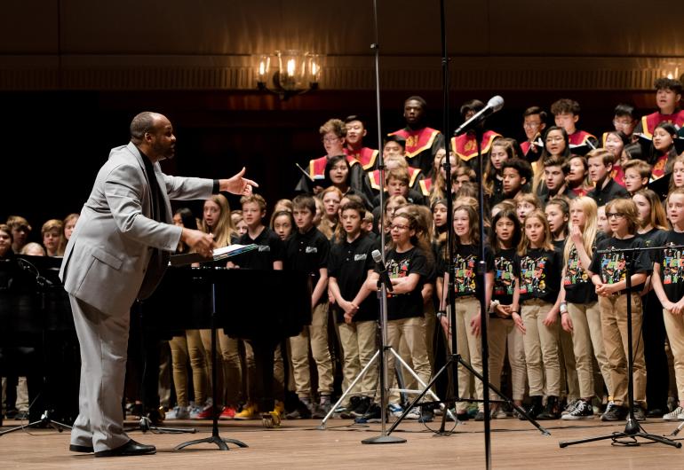 A person in a gray suit conducts a choir of children. 