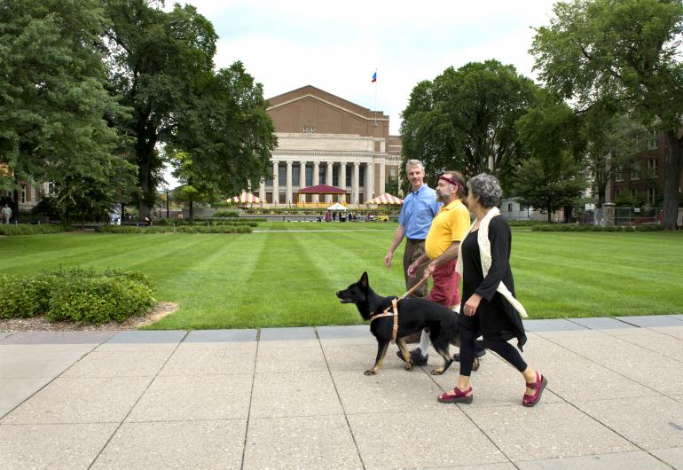 Three people walk with a dog in the University Mall. Northrop is in the background of the image.