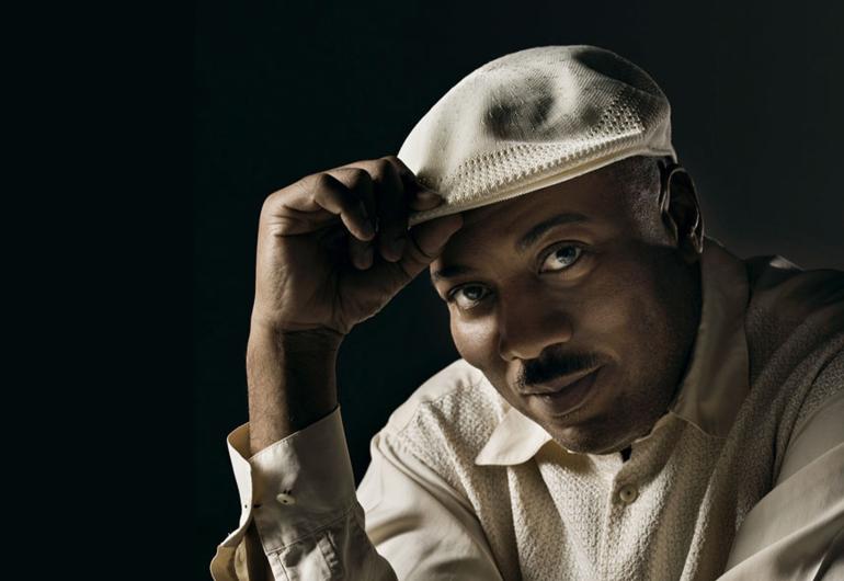 Alonzo King in a beige hat and shirt in front of a black background, looking into the camera.