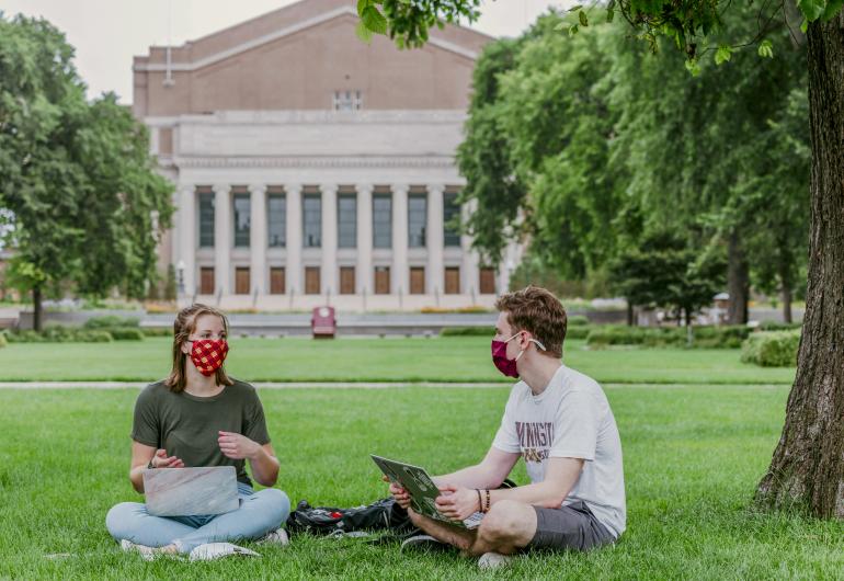 Students sit on lawn with Northrop in the background. Both students are wearing masks and sitting six feet apart.