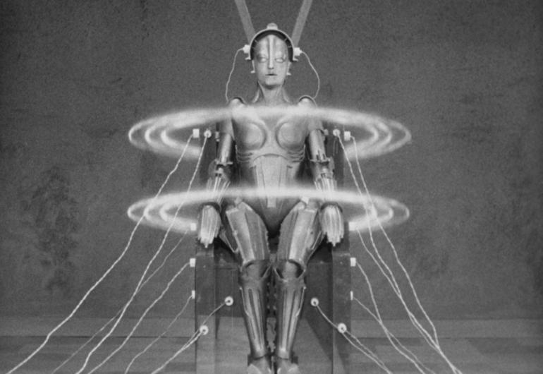 A human-like figure sits in a chair with wires connected to it. Two large circles float around the shoulders and waist of the figure. 