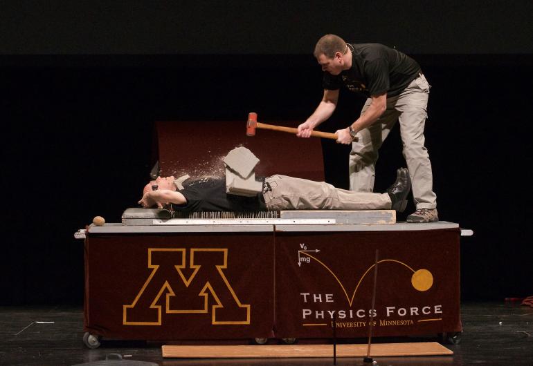 A person is laying on a bed of nails with another stands over him with a hammer, breaking a slab of concrete on his stomach.