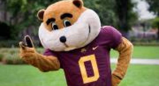 U of MN Mascot Goldie Gopher on the plaza in a football jersey giving the peace sign to the camera.