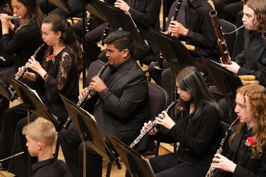 A row of young people seated and wearing black are holding a black and silver oboe instrument. The top is in the musicians' mouths and their hands are on the instrument.