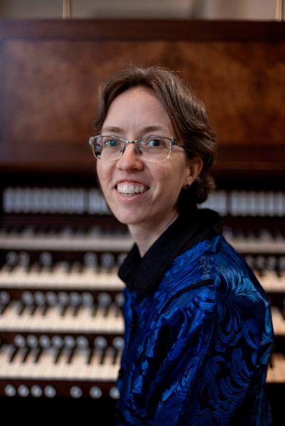 An organist wearing a deep blue button down with short, dark brown hair and glasses sits on a bench in front of a large, wooden organ console with their head turned to the left.