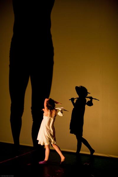 A dancer stands in front of an amber screen creating a shadow of a person wearing a hat and holding a stick with a bag attached.