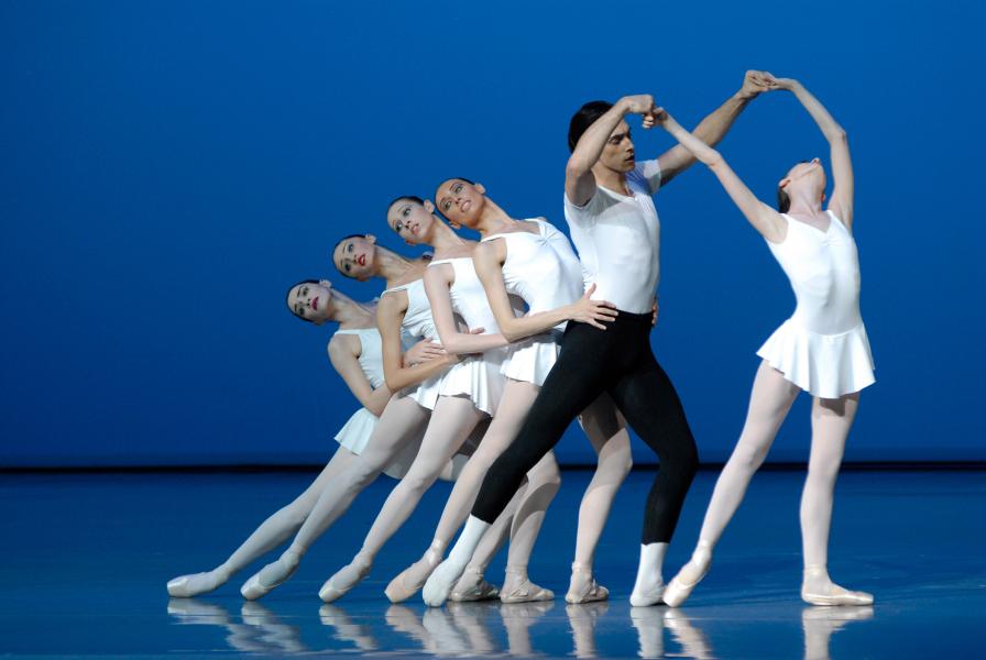 Five dancers stand in a line holding each other's waists. The dancer in the front of the line, wearing black pants and a white shirt, is holding a sixth dancer's arms above their head.