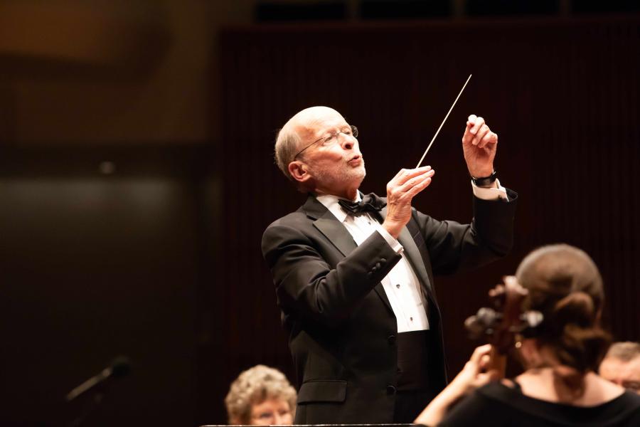 A conductor with grey hair, grey facial hair, and oval glasses, wears a black tuxedo and points a baton.