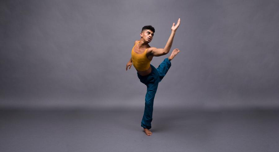 A dancer wearing a yellow top and blue pants appears in front of a gray backdrop. They stand on one leg with the other lifted up and behind them. One arm is raised and bent at the elbow while the other reaches behind them.