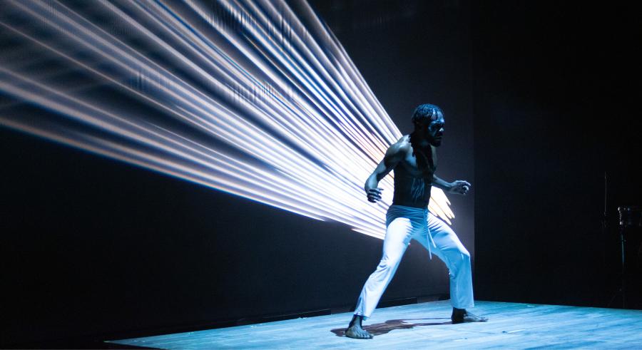 A dancer wearing white pants appears on a blue-lit stage, standing firmly with their feet planted and a projection of diagonal ray-like lines behind them. 