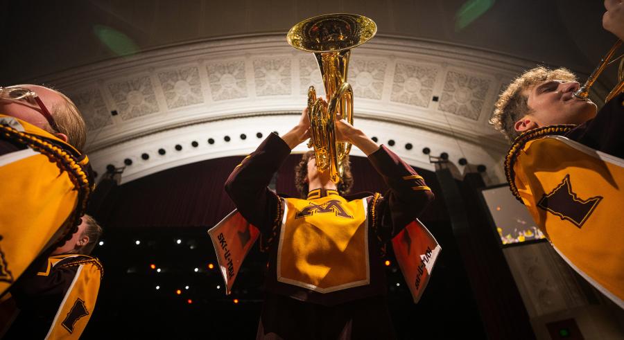 3 horn players wearing maroon and gold perform onstage. The camera is situated below one player, showing the underside of their trumpet.