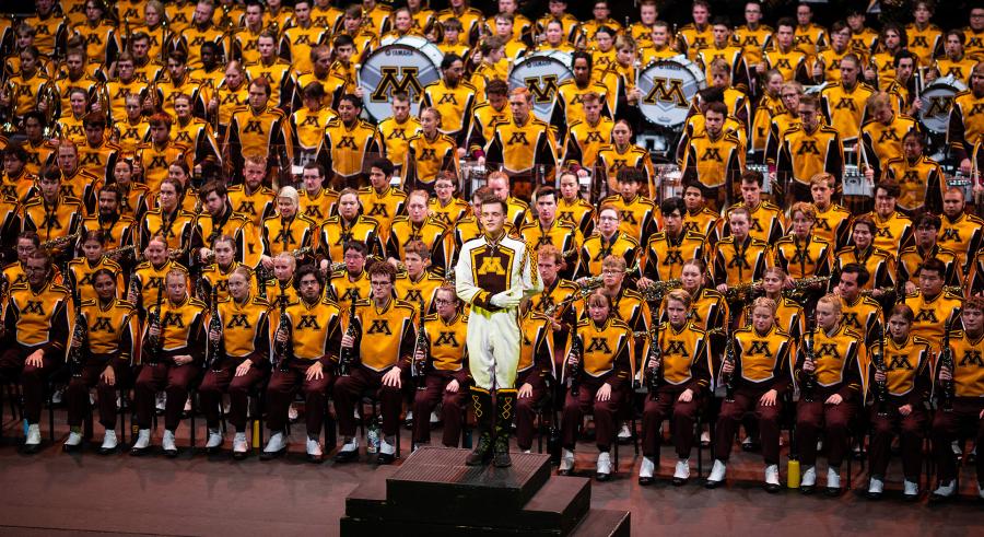 A marching band dressed in maroon and gold stands together onstage with the drum major slightly downstage on a podium. 3 large drums appear within the group, showcasing the University of Minnesota letter "M" logos.
