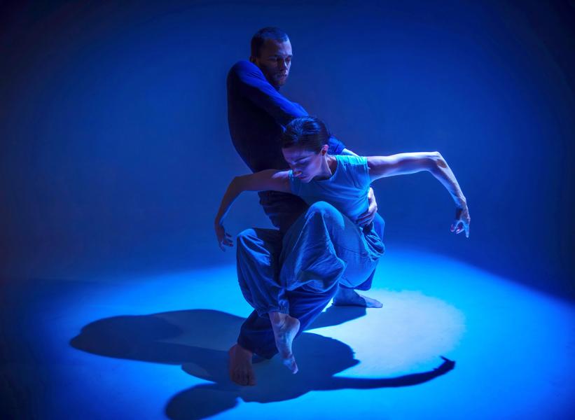 Couple in blue costumes, pose in a blue spotlight. Male dancer holds the waist of his partner as she is wrapped around his extended right leg, hovering above the floor with her arms bent out the side, elbows up and hands toward the floor.
