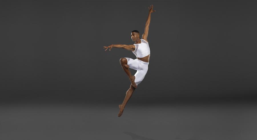 Male dancer in white leaps with toe to opposite knee and arms up and out