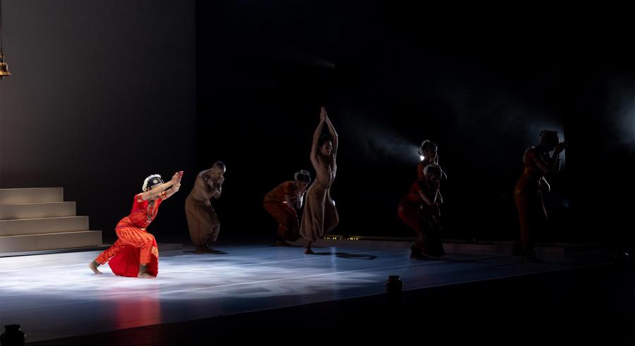 A dancer in red lunges in spotlight, others move in shadow.