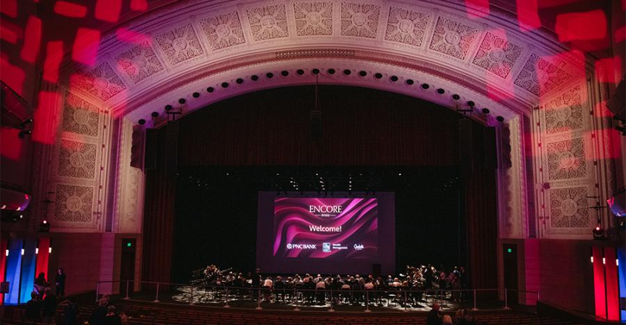 Inside Northrop’s auditorium, patrons sit along the stage facing a projector screen that reads “ENCORE 2022 Welcome!” with a purple background.White stone borders the opening for the stage and red rectangles are projected on the walls.