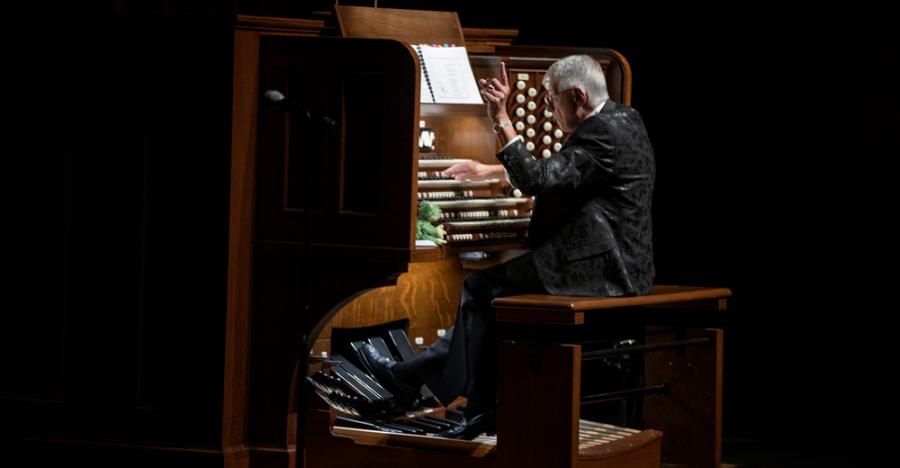 A dark stage shows the back of a man playing a large historic organ. He sits on the bench with his right hand on the organ keys, and his left hand in the air.