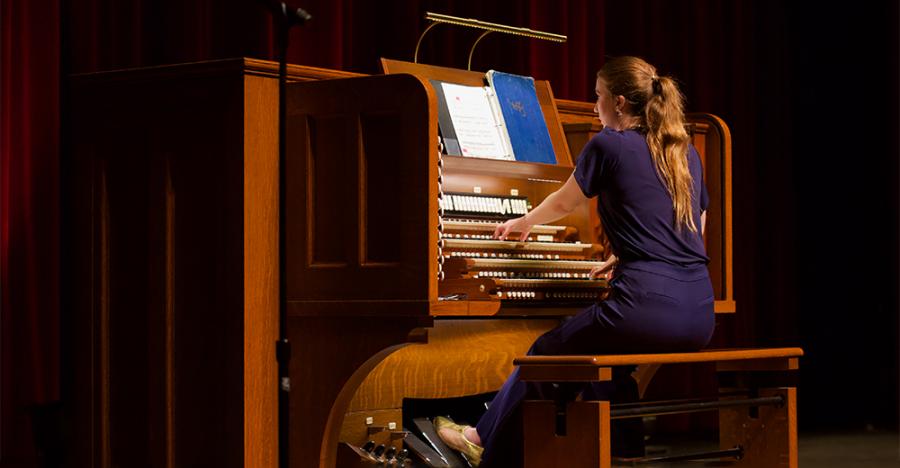 A person wearing a dark blue dress with light hair in a long ponytail, sits at a wooden Organ with their hands on the keys playing the instrument. 