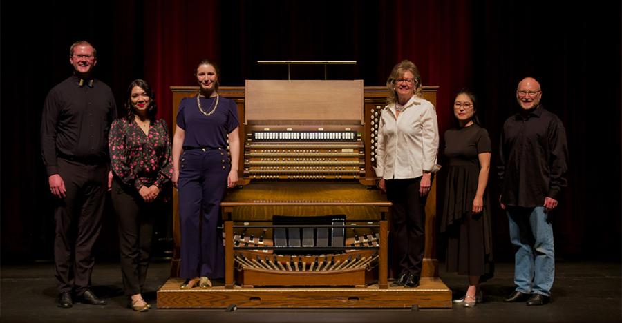 Six people stand around a wooden organ with three people on each side of the instrument. They all have their arms down and are smiling. The organ is on a black floor with red curtains behind it.
