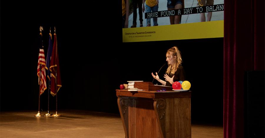 A female speaker stands at a wooden podium delivering a speech. There are three flags on the left side of the image and a projector screen in the back that shows the legs of three University students and a caption that reads, “Have found a way to balance.” 