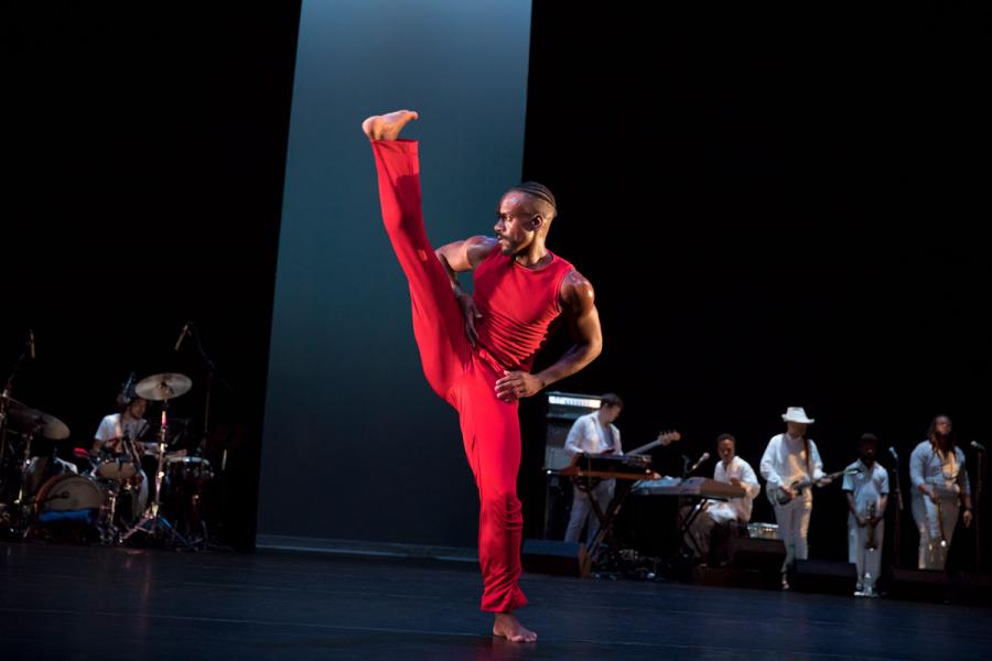 A male dancer wearing all red kicks his leg high on stage as a band plays in the background.