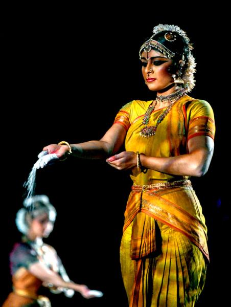 Female dancer in gold traditional Indian dress stands focusing as sand falls from her hand to the stage.