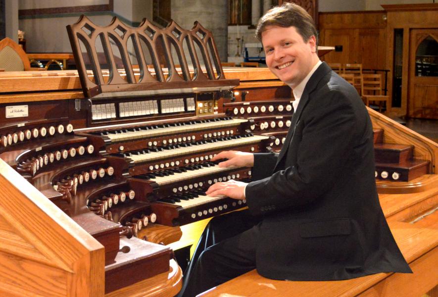Paul Jacobs sits at the organ looking to side, smiling at the camera