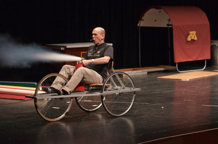 Man sits in a tri-wheeled chair holding a discharging fire extinguisher in his lap.