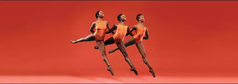 Three female dancers perform together in front of an orange background.