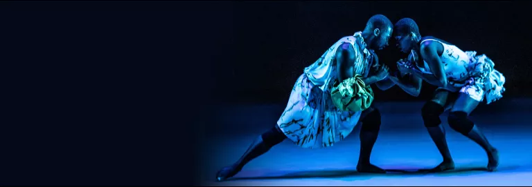 Two dancers, touching hands and leaning in towards eachother in a lunge position on a blue lit stage floor. Above hangs a neon green, glowing sphere of light.